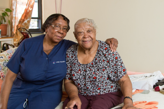 A VNS Health team member sits on a bed with her arm around an elderly woman.