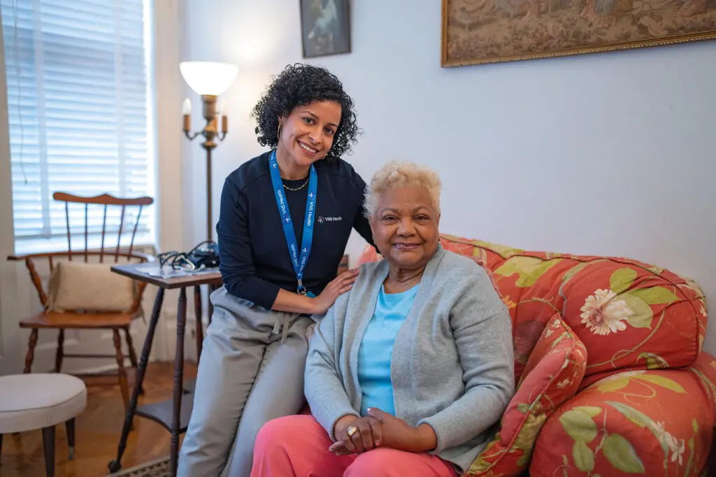 A female team member sits on the arm rest of an upholstered arm chair with her hand on the shoulder of a senior Black woman.