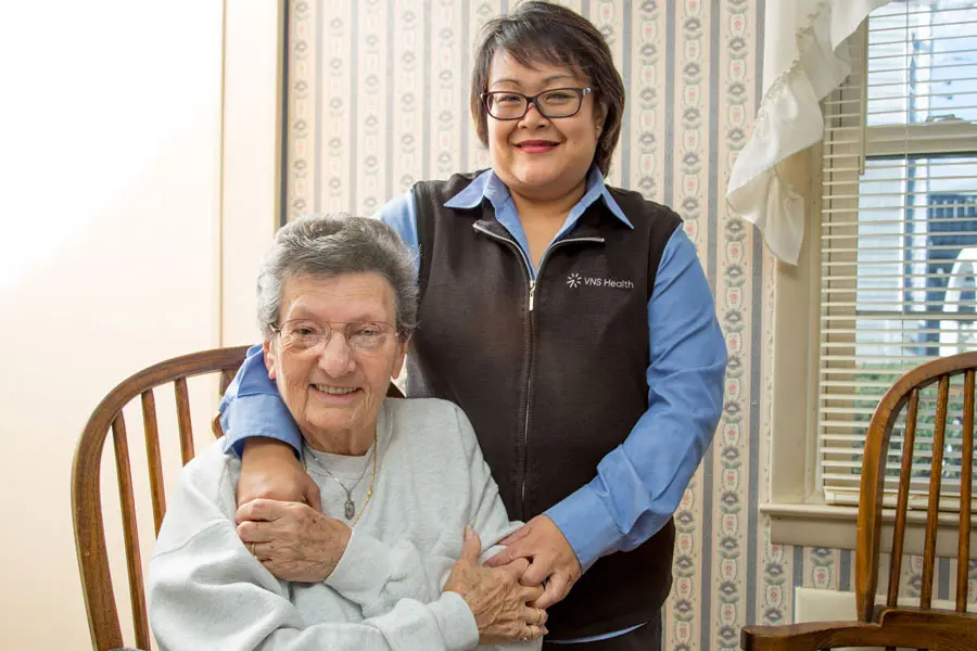 Nurse stands next to seated senior woman with their hands clasped on shoulder and by elbow