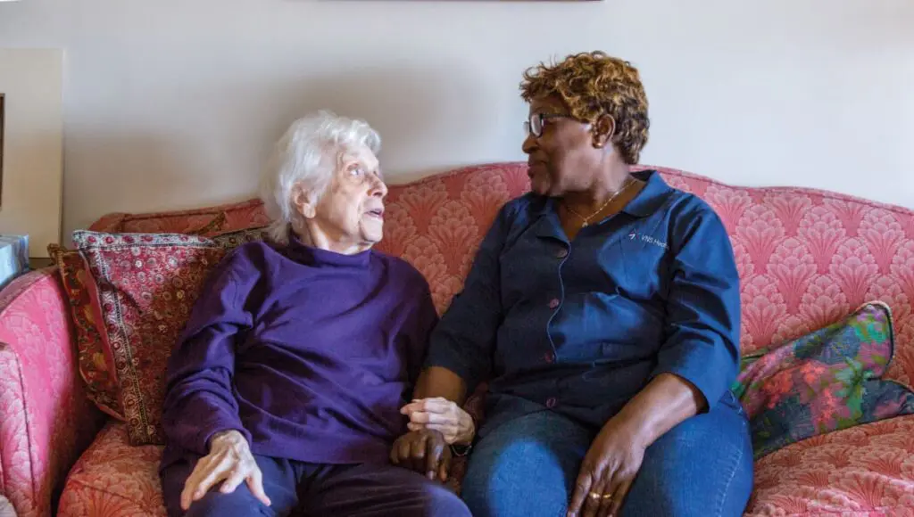 A VNS Health home health aide sits beside her patient receiving Dementia Care at home, their arms linked in a moment of care.