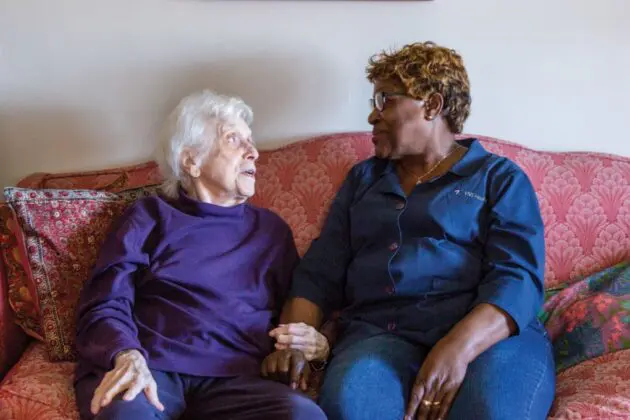A VNS Health home health aide sits beside her patient receiving Dementia Care at home, their arms linked in a moment of care.