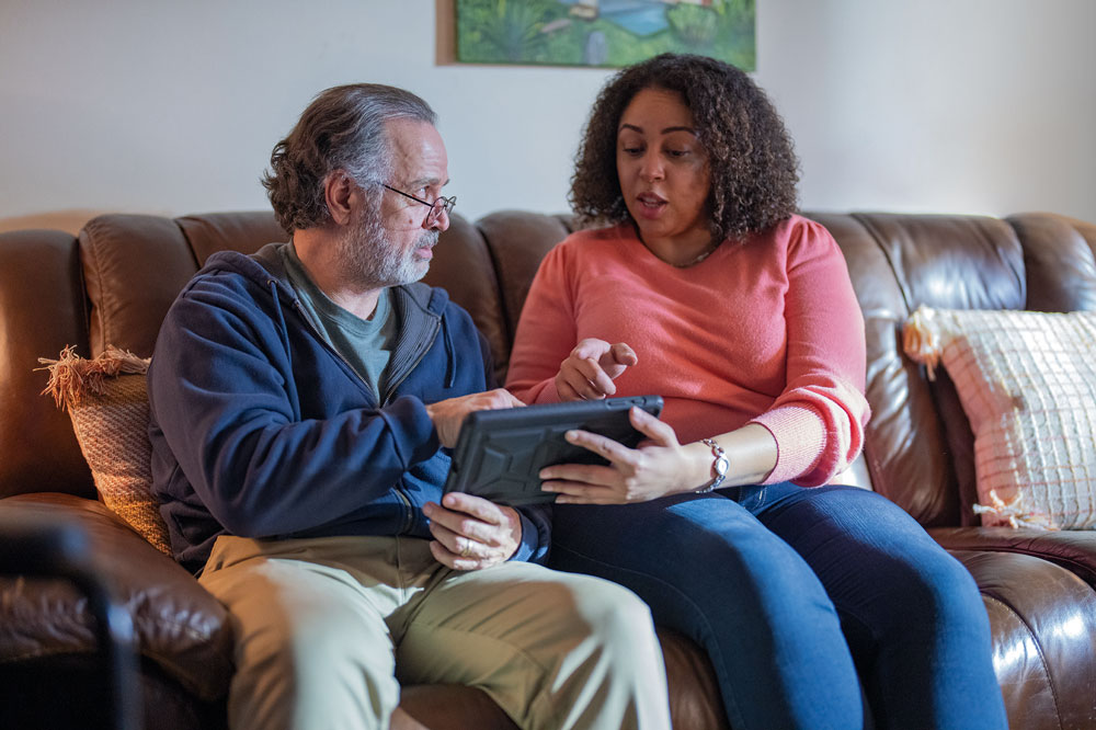 Adult daughter teaches her father how to use the electronic tablet as they sit on the couch.